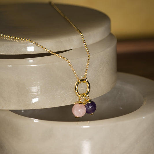 A gold chain necklace with a double-ring pendant holding two small beads, one light pink and one dark purple, displayed on a glossy light gray stone riser against a warm blurred background.