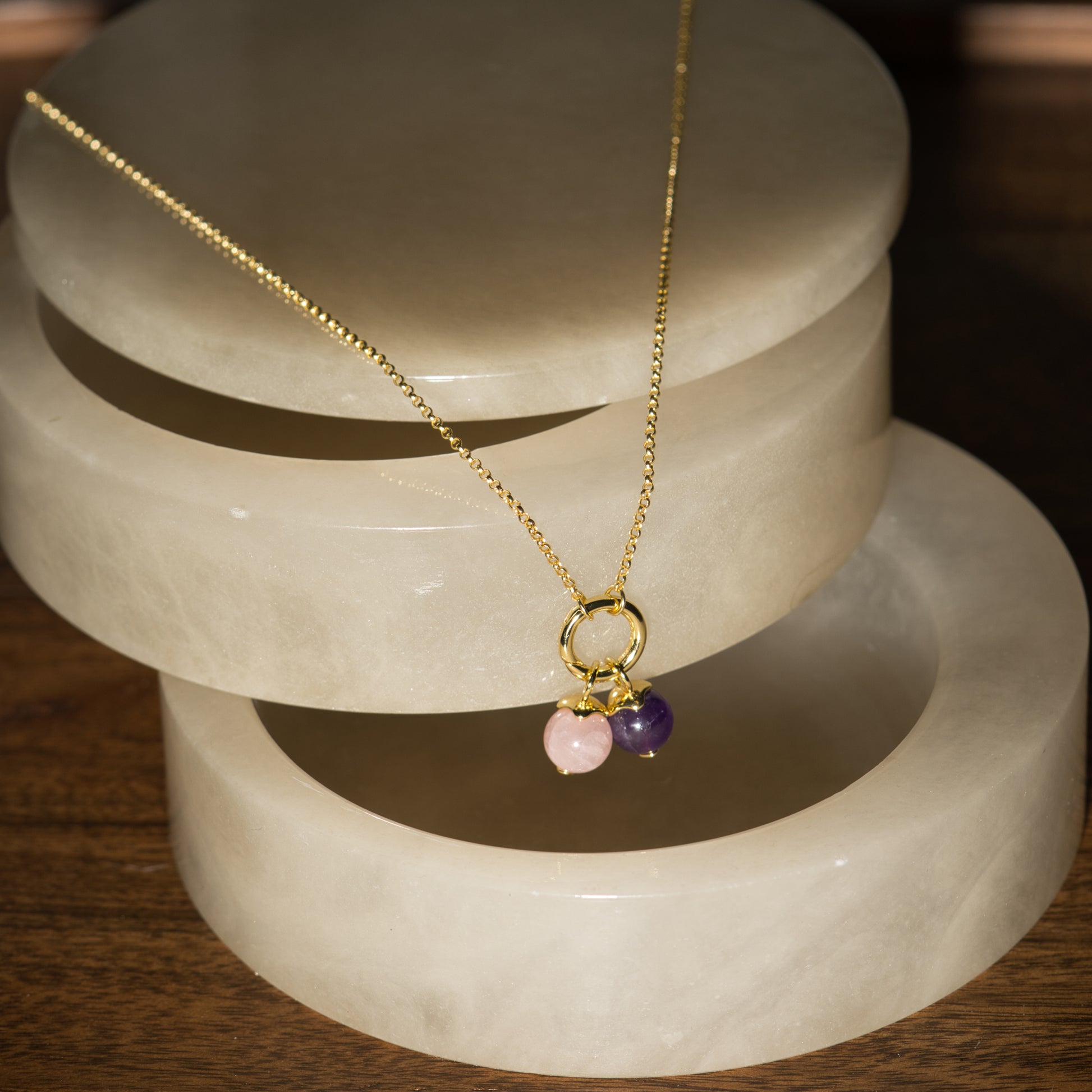A delicate gold chain necklace with a small gold ring pendant and two dangling beads, one light pink and one dark purple, displayed on a stacked white stone riser over a wooden surface.