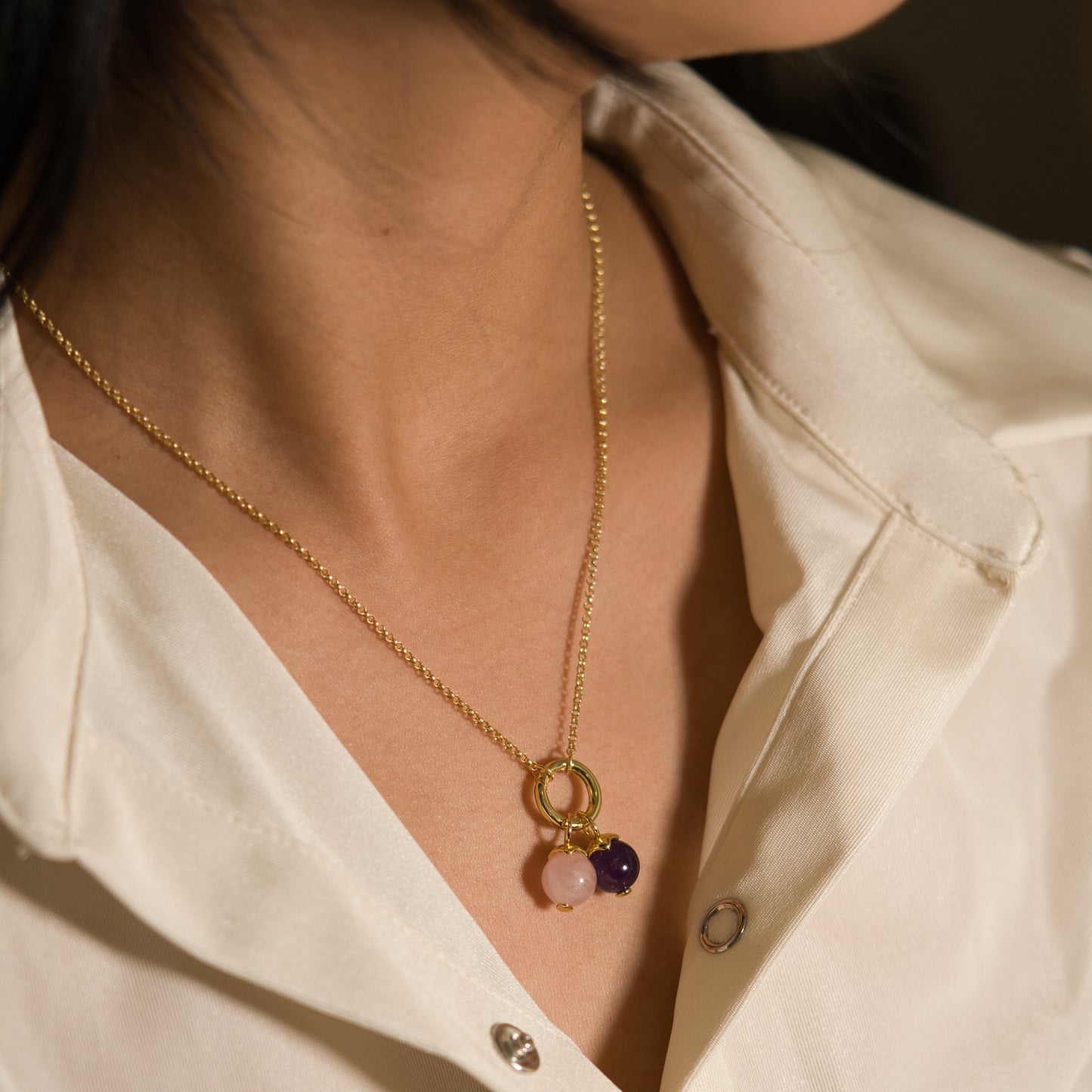 A close-up of a woman wearing a delicate gold chain necklace with a pendant featuring a gold ring and two small gemstone charms, one light pink and one dark purple, styled with a white collared shirt.