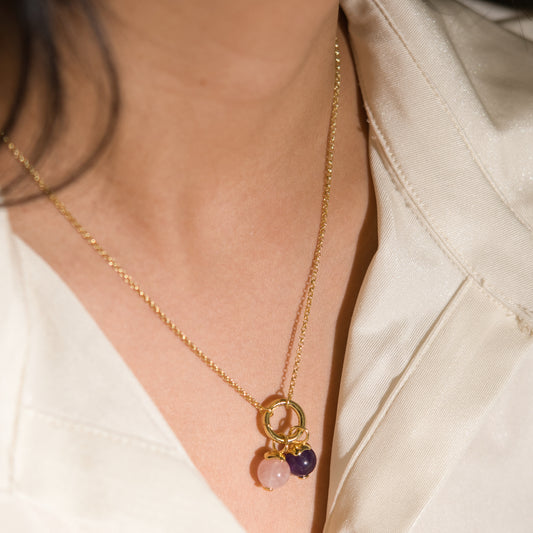 A close-up of a person wearing a delicate gold chain necklace with a double-ring pendant and two small hanging beads, one light pink and one dark purple, against a cream satin blouse.